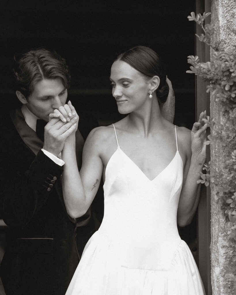 Groom in velvet tuxedo kisses bride’s hand as she smiles in Danielle Frankel gown at Monteverdi Tuscany.