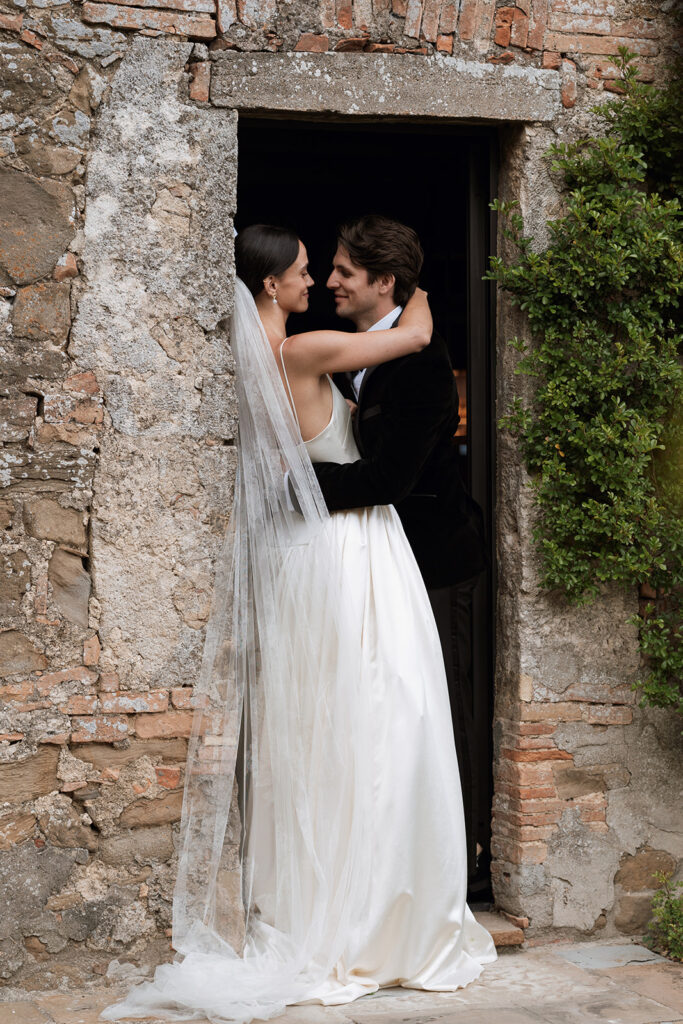 Bride in Danielle Frankel gown embraces groom in velvet tuxedo in a stone doorway at Monteverdi Tuscany.