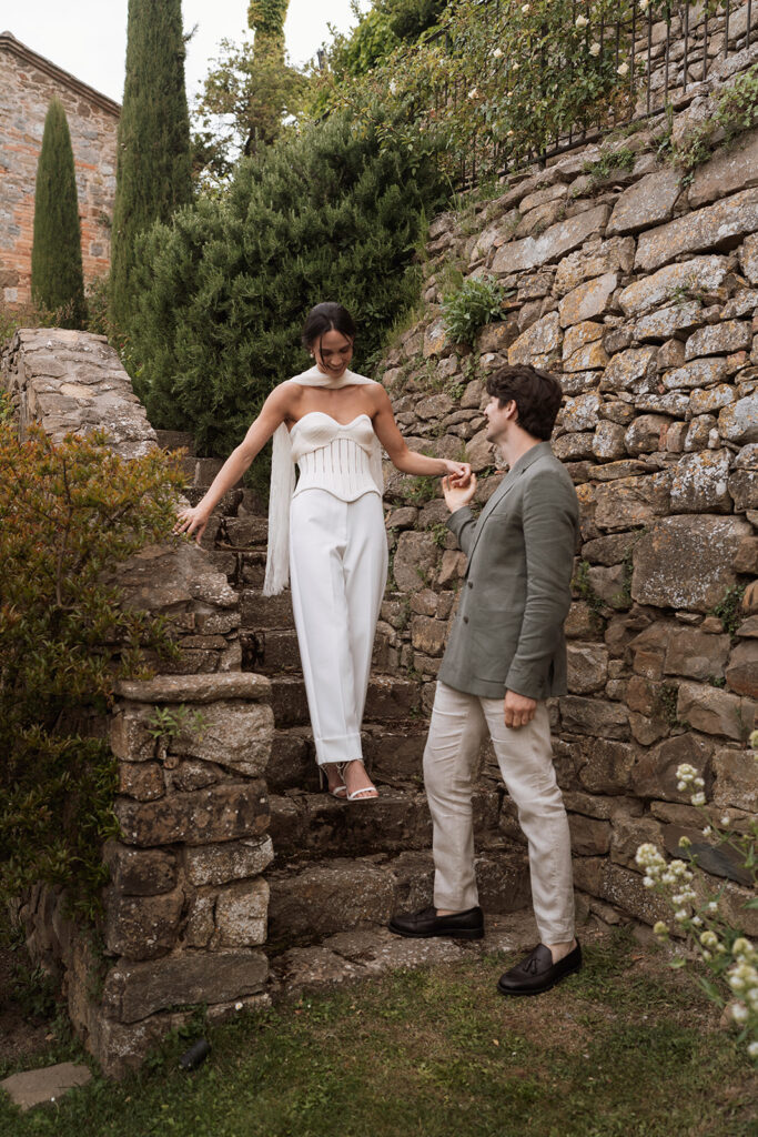 Bride wearing a Danielle Frankel corset descends stone steps at Monteverdi Tuscany while holding her groom’s hand.