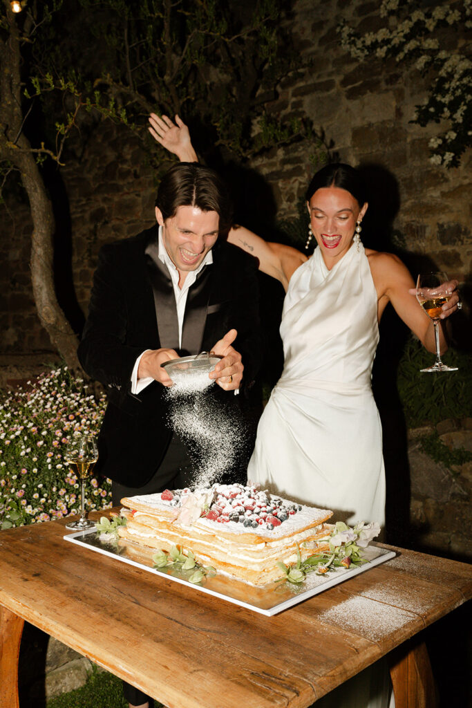 Bride and groom celebrate while dusting powdered sugar over millefoglie cake at Monteverdi Tuscany.
Caption: