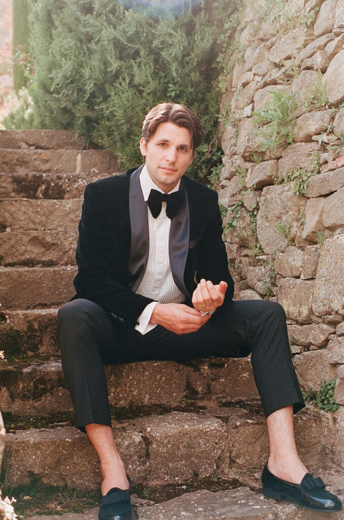 Groom in a black velvet tuxedo sitting on rustic stone steps at Monteverdi Tuscany during golden hour.