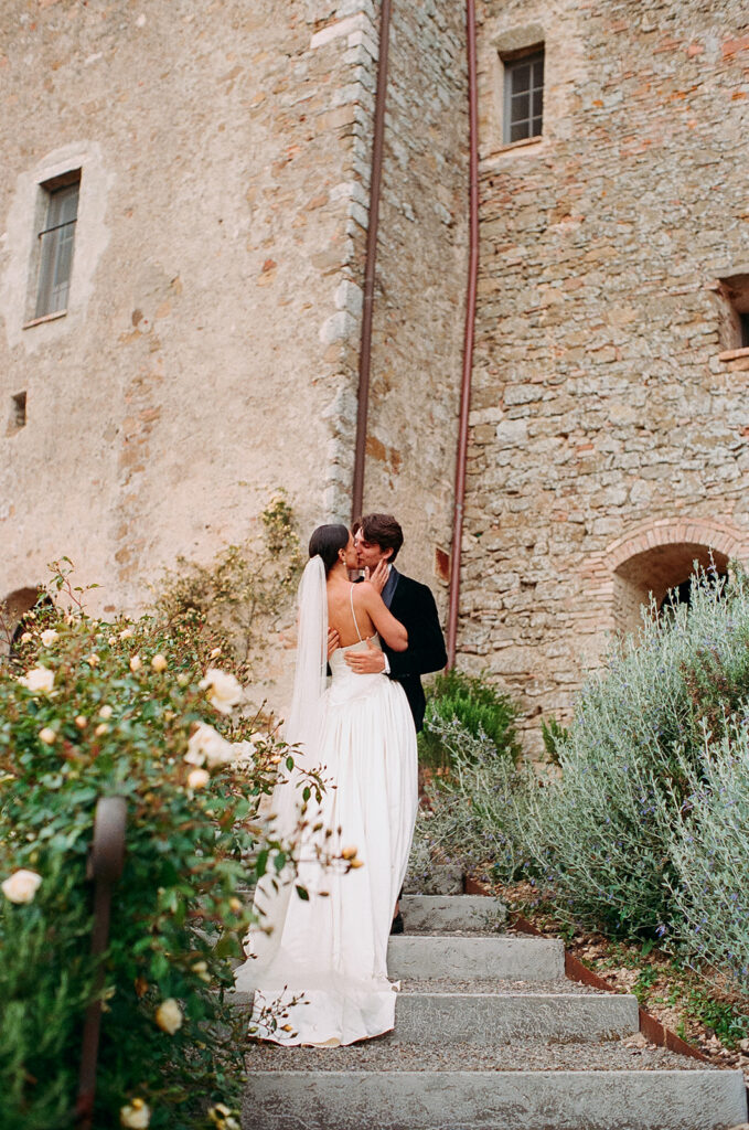 Bride in Danielle Frankel gown and Jane Rhyan The Collection veil shares a kiss with groom in black velvet tuxedo on stone steps at Monteverdi Tuscany.