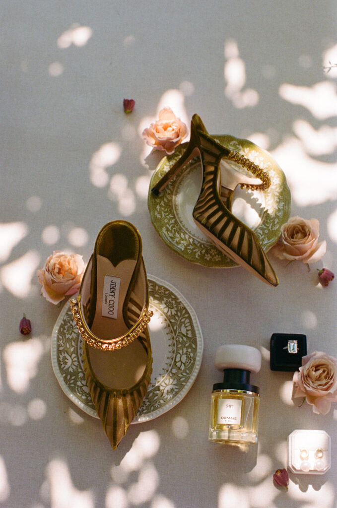 Editorial flat lay of bride’s wedding details featuring gold Jimmy Choo heels, vintage green china, Ormaie perfume, and diamond ring at Monteverdi Tuscany.