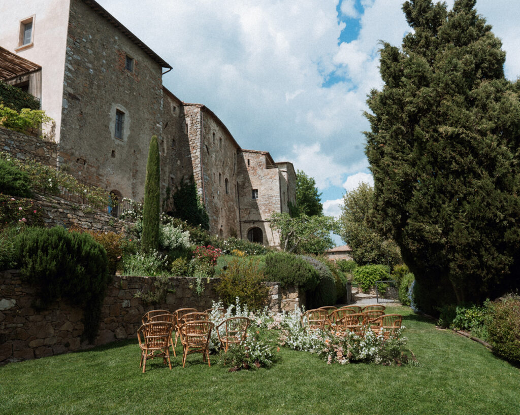 Outdoor wedding ceremony setup at Monteverdi Tuscany with rattan chairs and garden florals against ancient stone architecture.