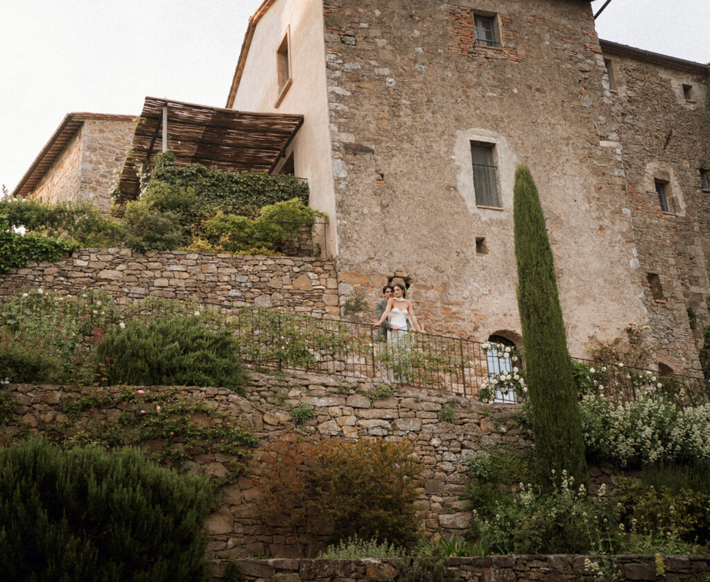 Bride and groom standing on a terrace overlooking Monteverdi Tuscany’s historic stone walls and cascading gardens.