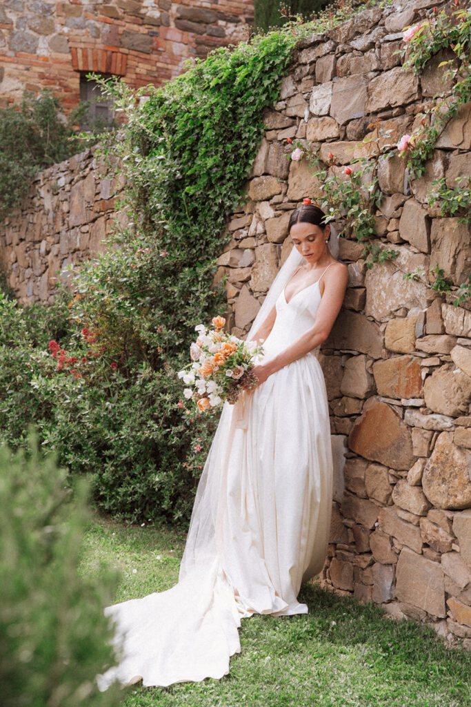 Bride in a Danielle Frankel gown and Jane Rhyan The Collection veil, standing against a rustic stone wall at Monteverdi Tuscany.