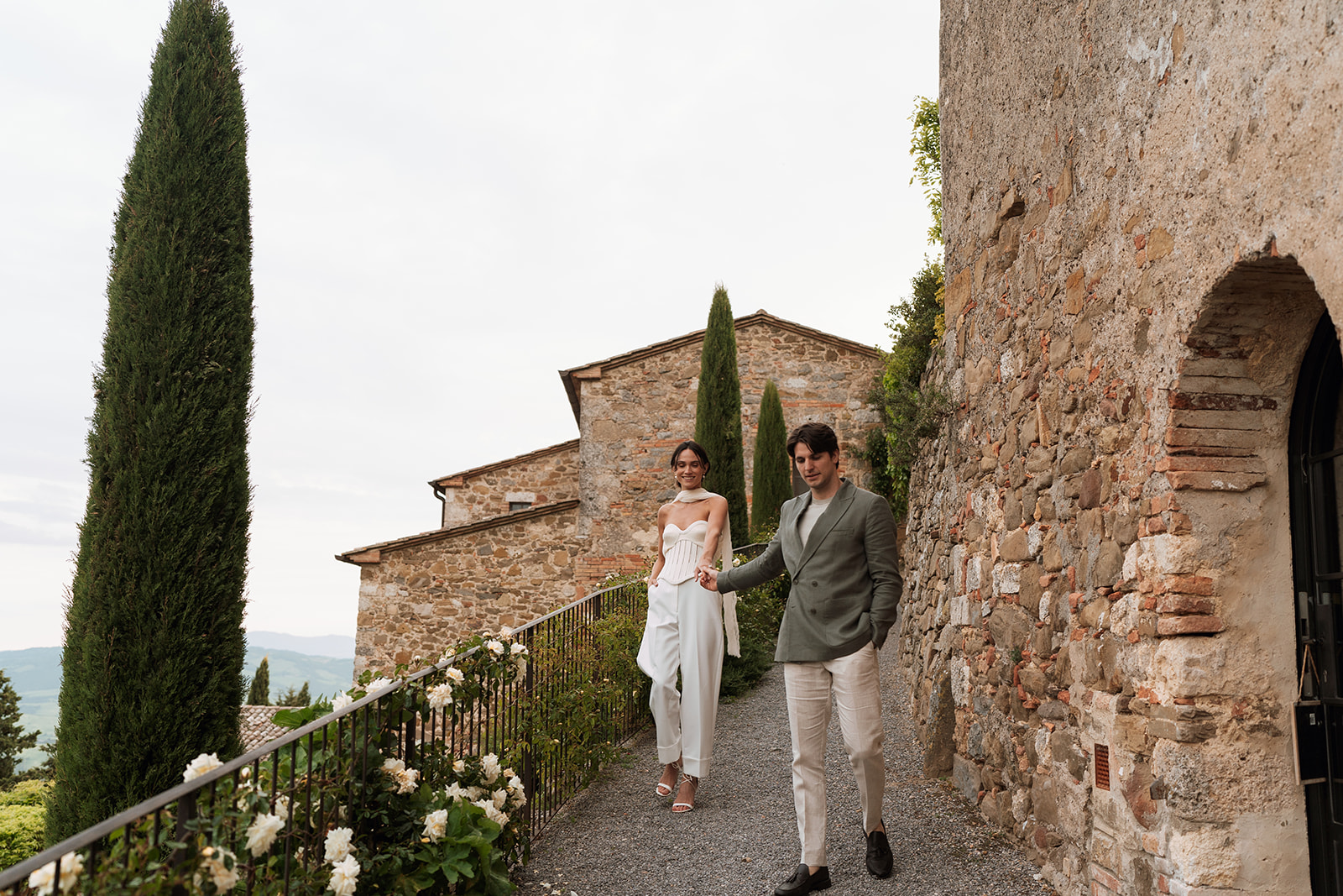 Bride and groom walking hand in hand along the cobblestone pathway at Monteverdi Tuscany, surrounded by cypress trees and stone villas.