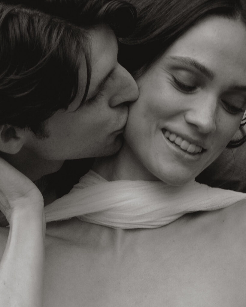 Intimate black and white close-up of groom kissing bride’s cheek at Monteverdi Tuscany, her scarf softly framing her shoulders.