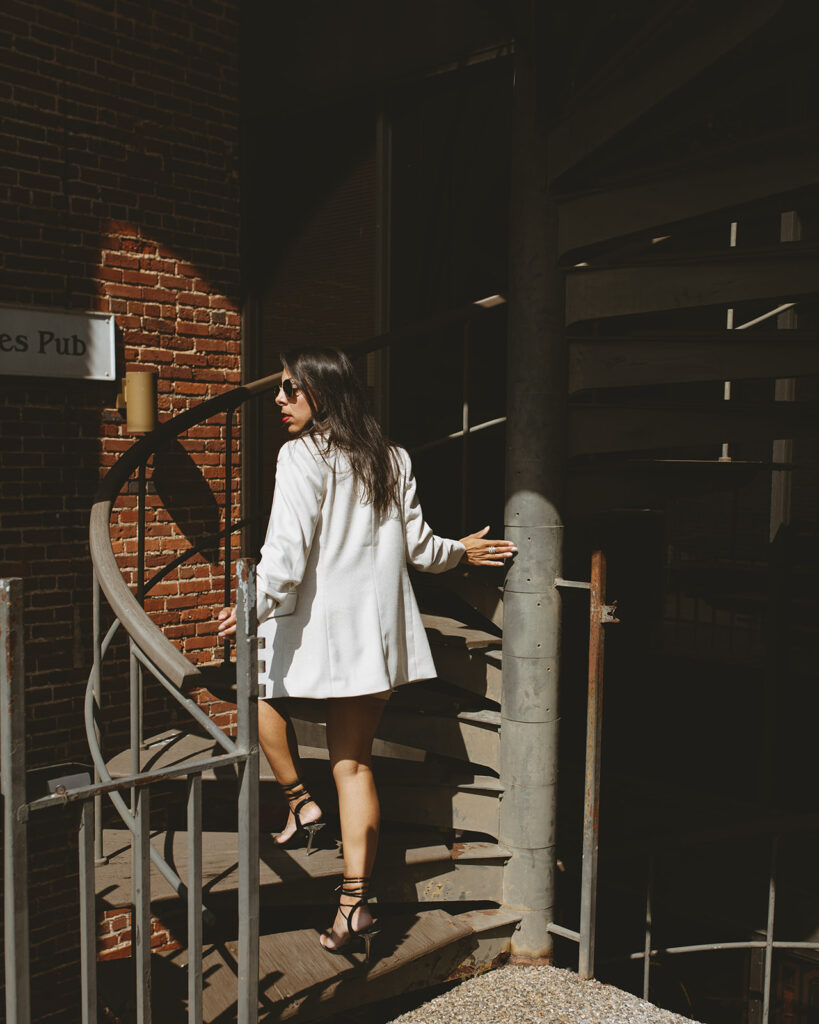 Woman walking up an outdoor spiral staircase in sunlight, wearing a white blazer and heels, beside a brick wall.