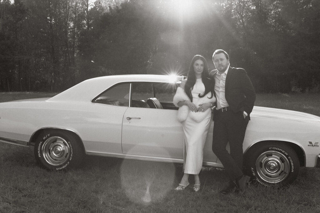 A black-and-white photo of an elegant couple leaning against a vintage yellow Chevrolet Chevelle in a sunlit field, captured with soft film tones and cinematic light flares.