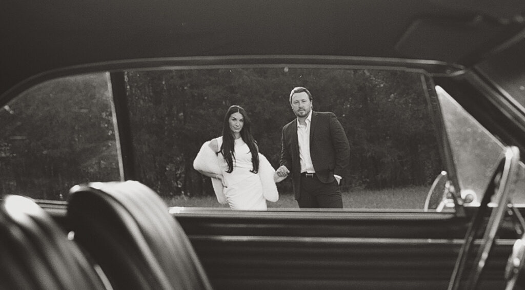 Couple standing outside a vintage car, framed through the open window, captured in black and white during an editorial film-inspired session near Charlotte.