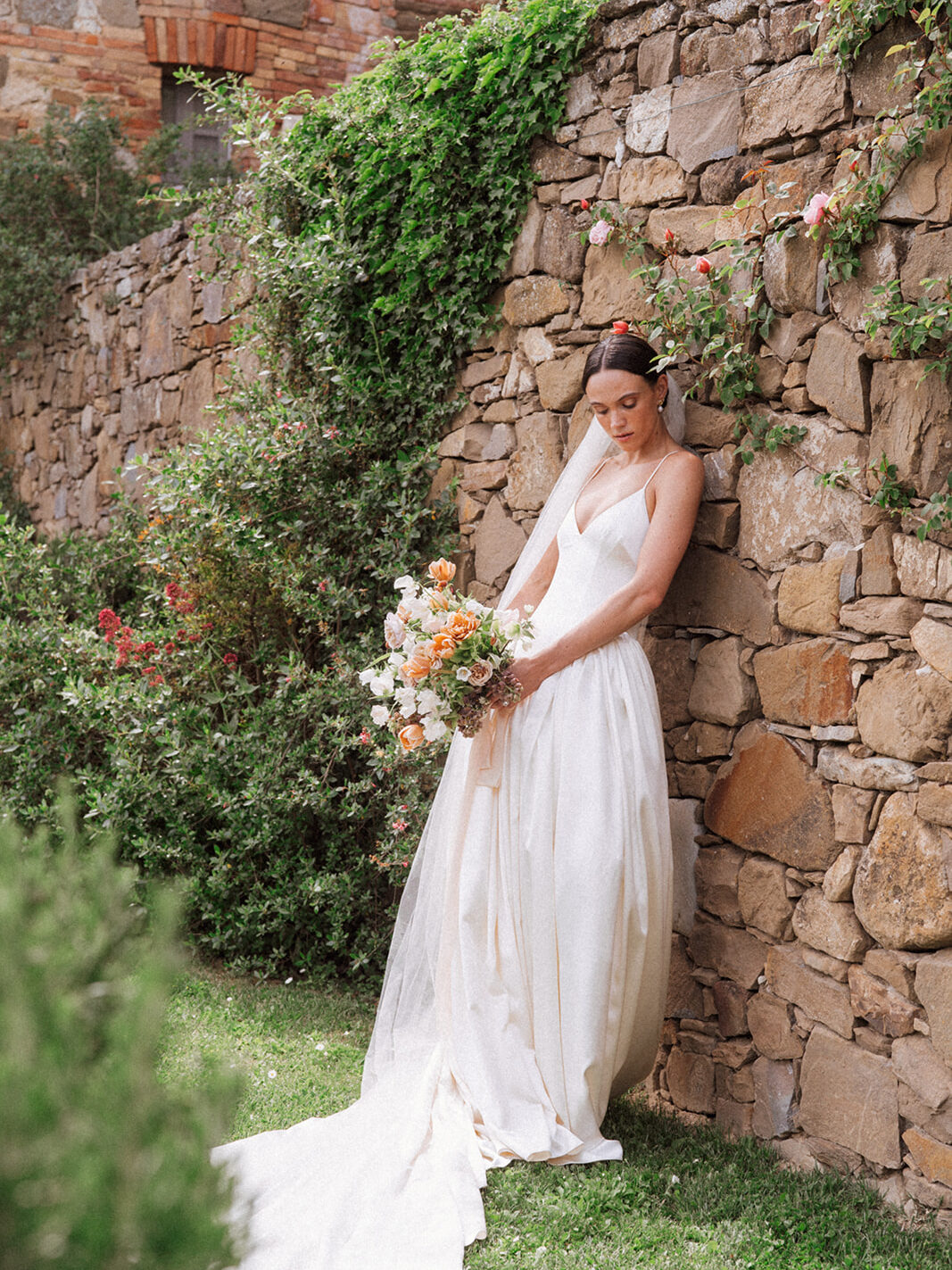 A Timeless Monteverdi Tuscany Wedding Bride in a Danielle Frankel gown and Jane Rhyan The Collection veil, standing against a rustic stone wall at Monteverdi Tuscany.